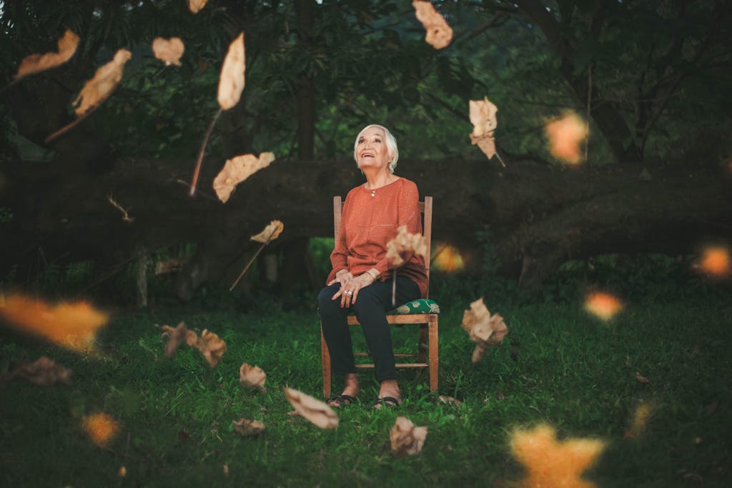 An elderly woman sits outdoors enjoying the falling autumn leaves, capturing a serene moment.